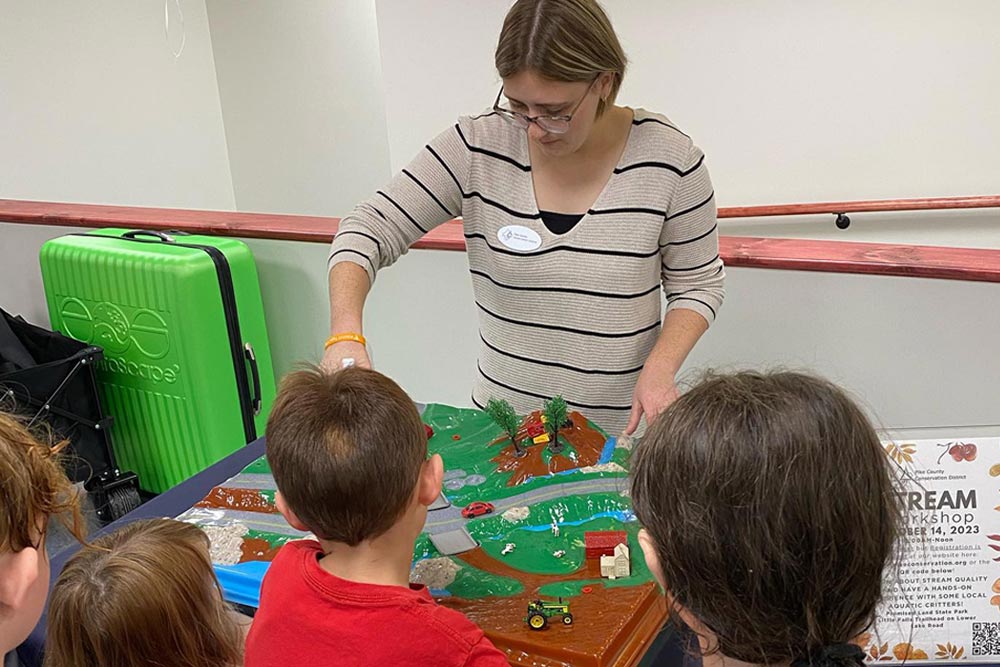 A group of children gather around a table with an Enviroscape watershed model and an educator demonstrating how pollution travels through the model