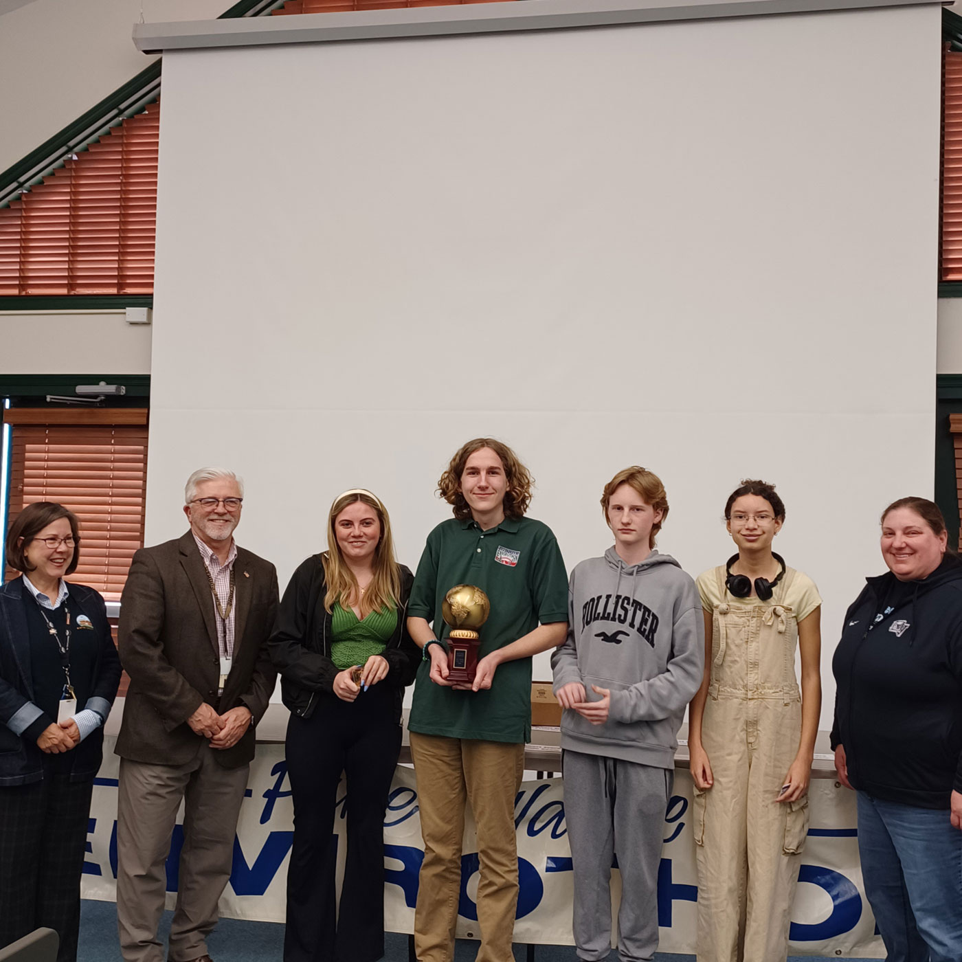 A group of high school students, their teacher, and two county commissioners stand in front of an Envirothon banner, the middle student holding a trophy