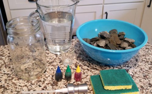 The supplies needed for an at-home groundwater model set on a counter, including a bowl of rocks, two sponges, three colors of food dye, a pitcher of water, a glass jar, and a hand soap dispenser