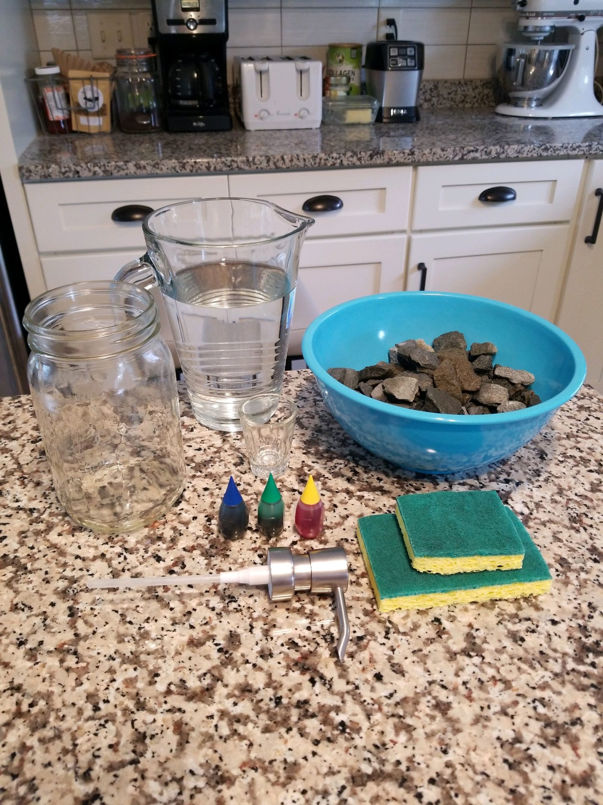 The supplies needed for an at-home groundwater model set on a counter, including a bowl of rocks, two sponges, three colors of food dye, a pitcher of water, a glass jar, and a hand soap dispenser