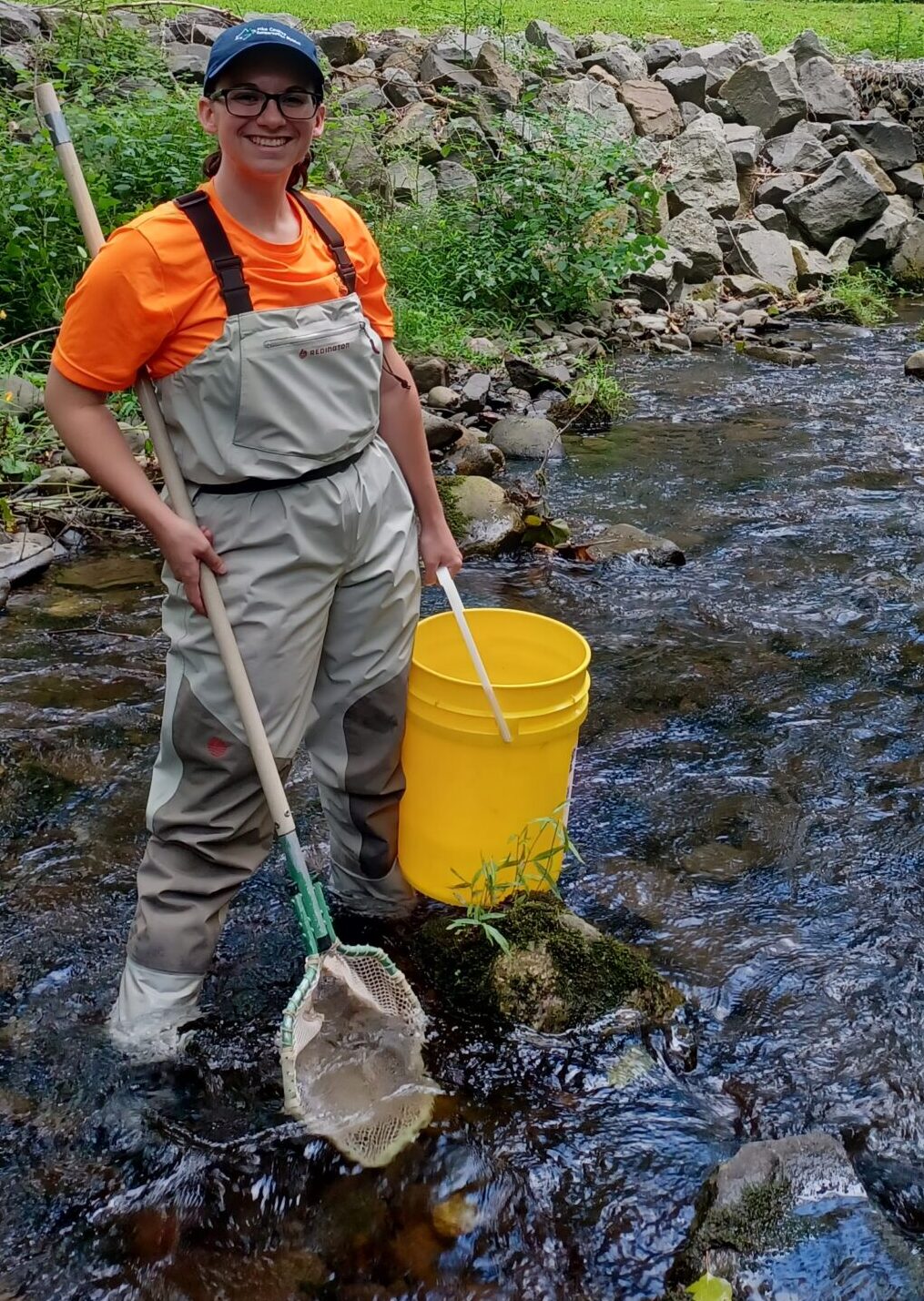 Watershed Specialist stands in a stream holding a net and a bucket