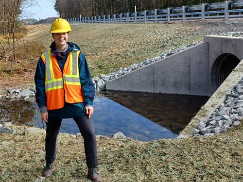 Resource Conservationist stands in a hard hat and safety vest next to a road culvert